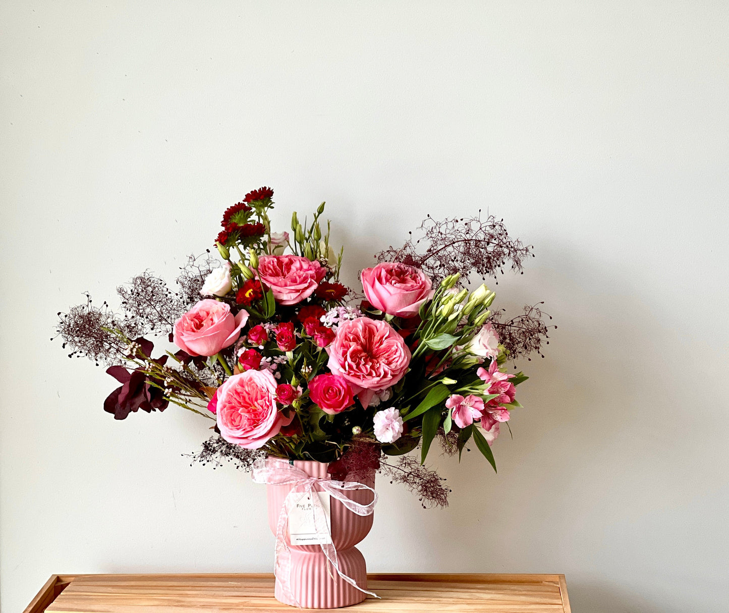 Elegant pink vase arrangement with large pink garden roses, red spray roses, lisianthus, and delicate burgundy foliage, tied with a ribbon from Five Petals Florist.