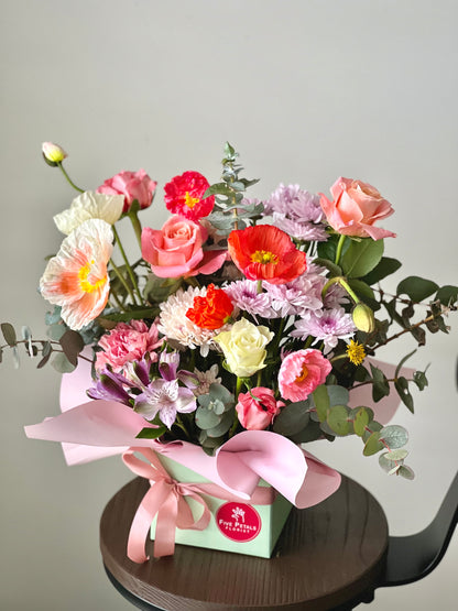Vibrant flower arrangement in a pastel green box with pink ribbon, featuring poppies, roses, eucalyptus, and delicate blooms from Five Petals Florist.