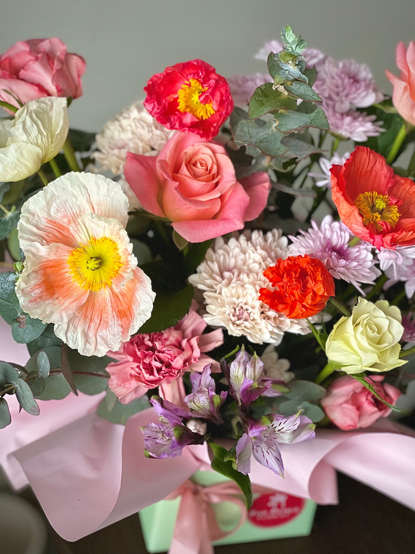 Vibrant flower arrangement in a pastel green box with pink ribbon, featuring poppies, roses, eucalyptus, and delicate blooms from Five Petals Florist.
