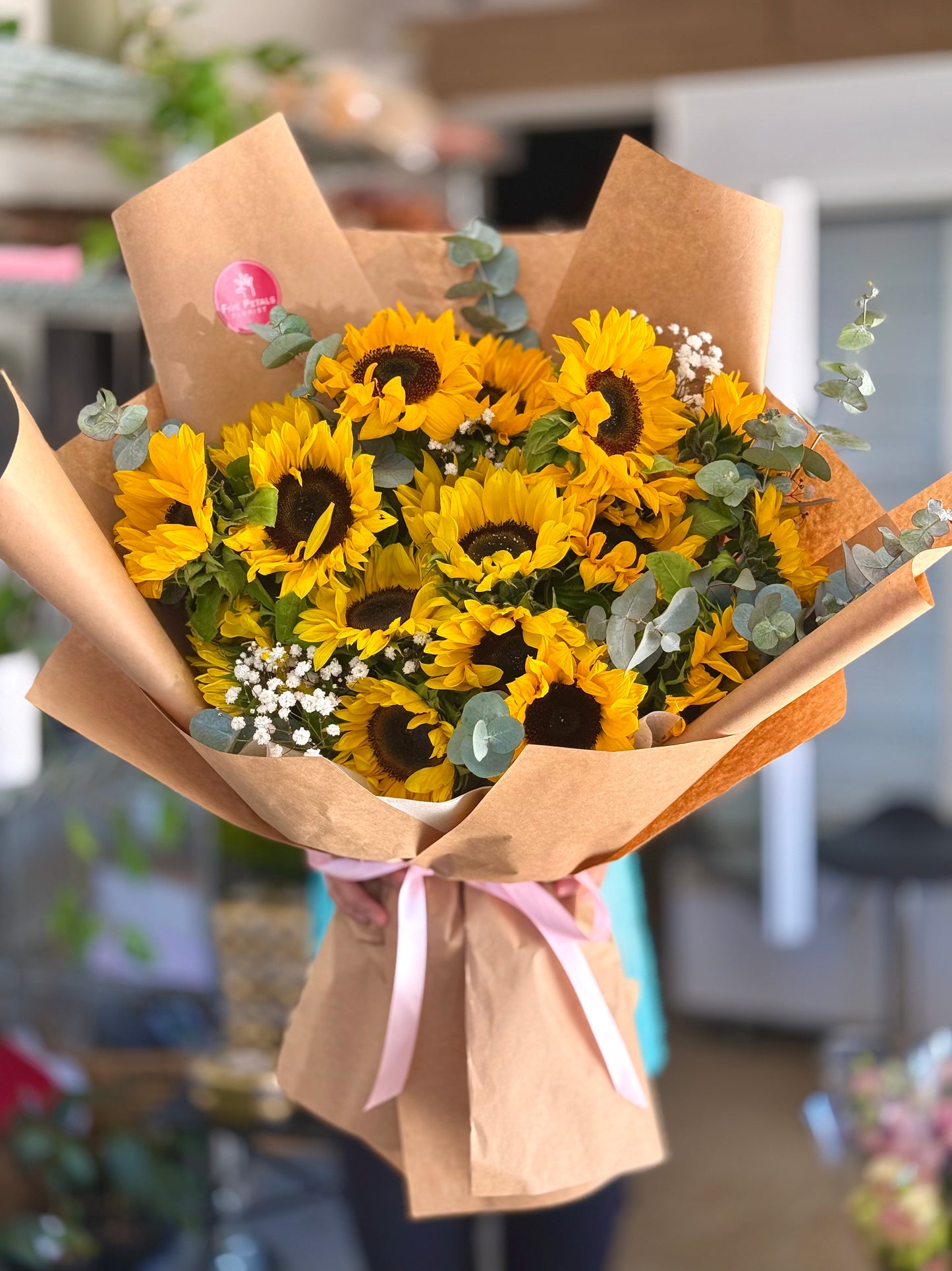 Bouquet of sunflowers wrapped in brown paper with a pink ribbon, by Five Petals Florist.