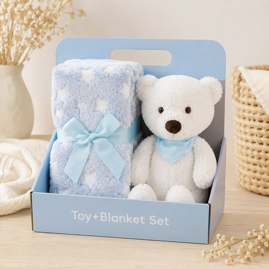 A baby gift set in a light blue box labeled 'Toy + Blanket Set', containing a white plush teddy bear with a light blue bow and a light blue blanket with white star patterns and a matching bow.